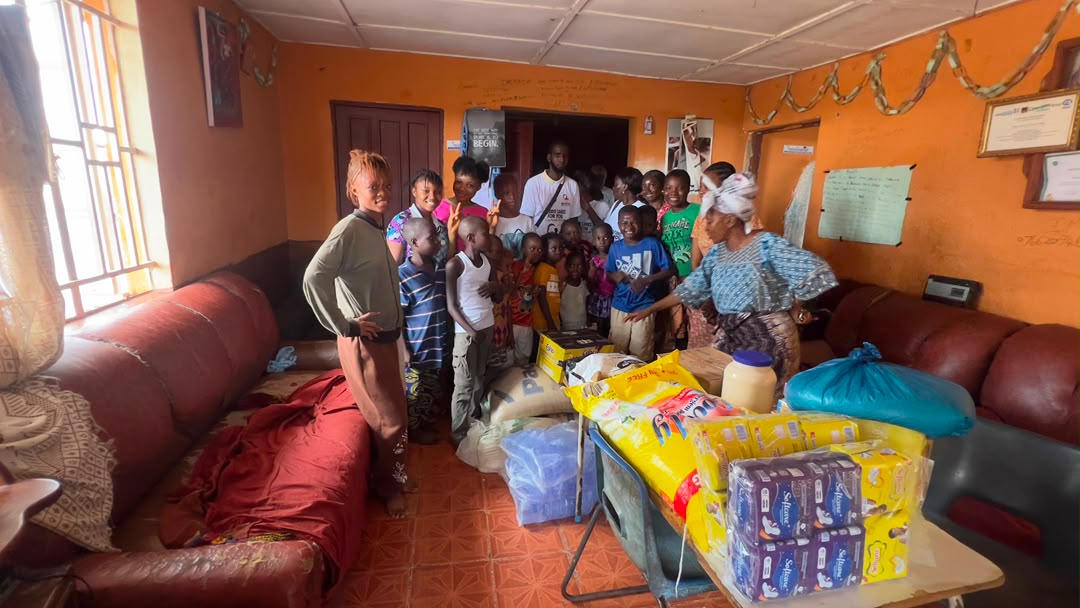 Children and their carer stand in a room beside donated rice, flour and household basics